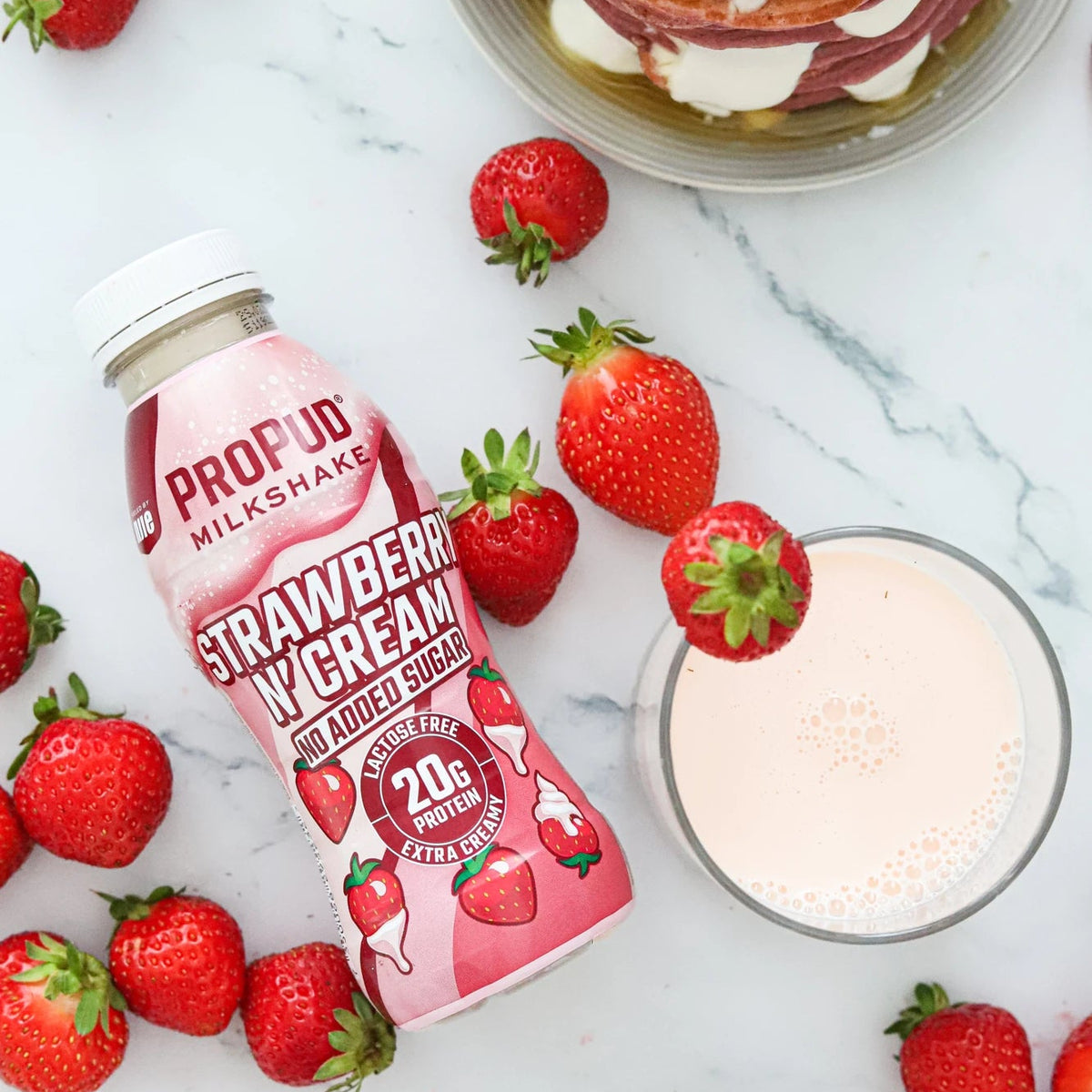 Two bottles of Profud Milkshake Strawberry Creamer with red strawberries and a glass of the pink drink, styled on a white marble surface with a plate of pancakes in the background.